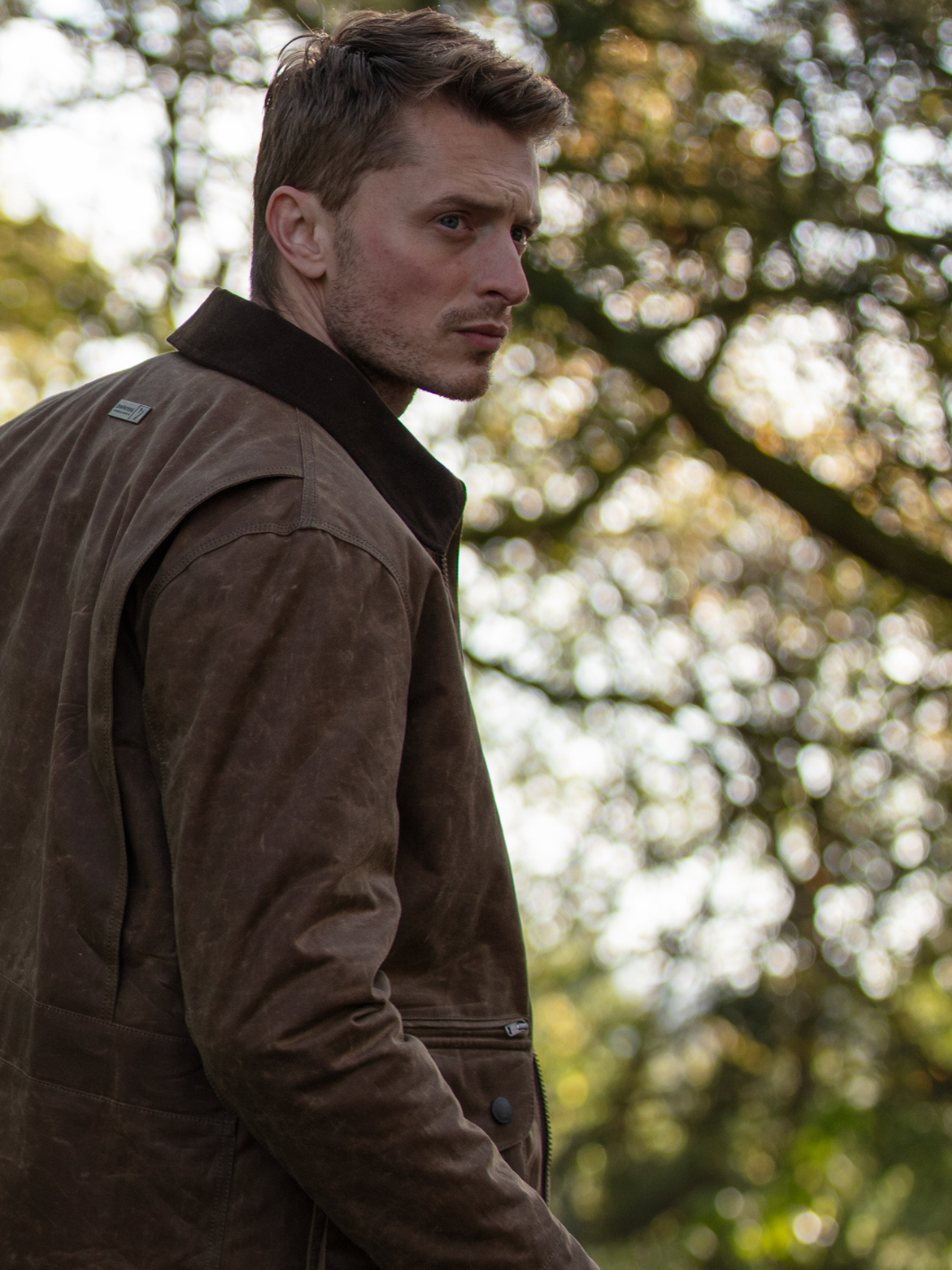 Hinterland Jacket in heavy tan waxed cotton with a desert finish, featuring a hip-length design, oversized fit, quilted brown lining, dark brown moleskin trims, and antique brass plate. Includes 2 front pockets, zipped pocket, inner wallet pocket, and YKK zip with storm flap. Limited edition, part of Partridge England's range.