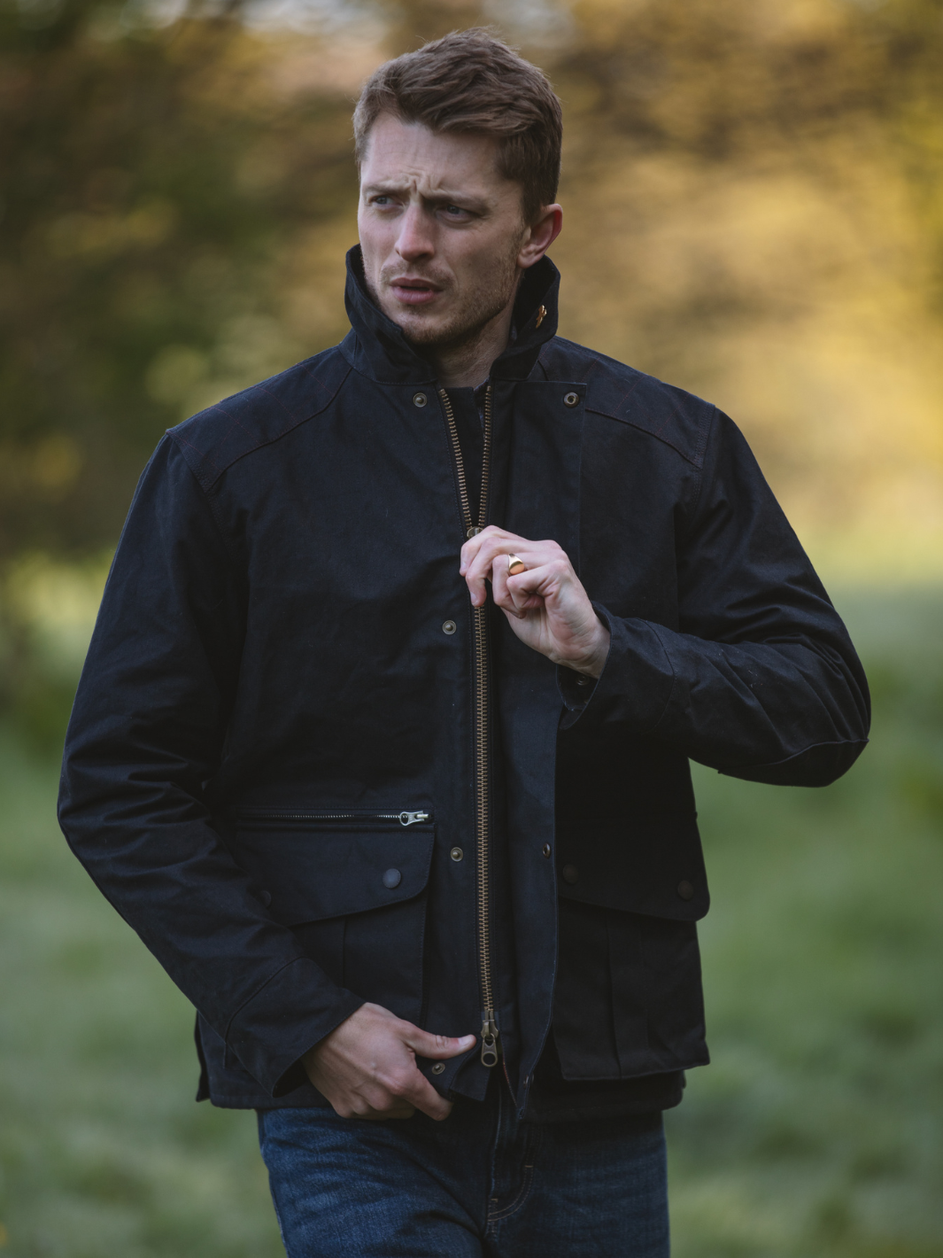 Hinterland Hybrid Jacket made of black dry waxed canvas with quilted shoulder and elbow patches, black moleskin trims, and red and burgundy check lining. Features include a hip-length design, oversized fit, action back, antique brass plate, and YKK zip with storm flap. Limited edition, part of the Partridge England range.