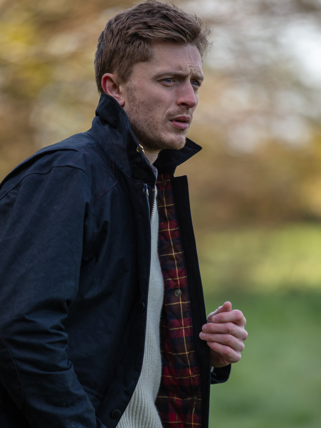 Hinterland Hybrid Jacket made of black dry waxed canvas with quilted shoulder and elbow patches, black moleskin trims, and red and burgundy check lining. Features include a hip-length design, oversized fit, action back, antique brass plate, and YKK zip with storm flap. Limited edition, part of the Partridge England range.