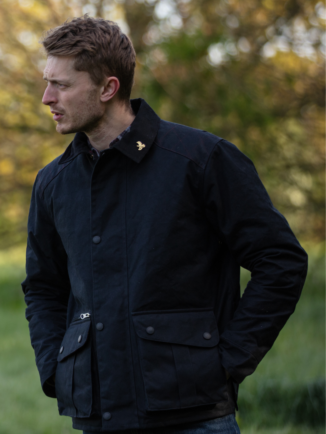 Hinterland Hybrid Jacket made of black dry waxed canvas with quilted shoulder and elbow patches, black moleskin trims, and red and burgundy check lining. Features include a hip-length design, oversized fit, action back, antique brass plate, and YKK zip with storm flap. Limited edition, part of the Partridge England range.
