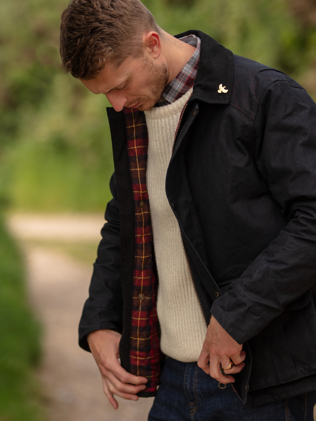 Hinterland Hybrid Jacket made of black dry waxed canvas with quilted shoulder and elbow patches, black moleskin trims, and red and burgundy check lining. Features include a hip-length design, oversized fit, action back, antique brass plate, and YKK zip with storm flap. Limited edition, part of the Partridge England range.
