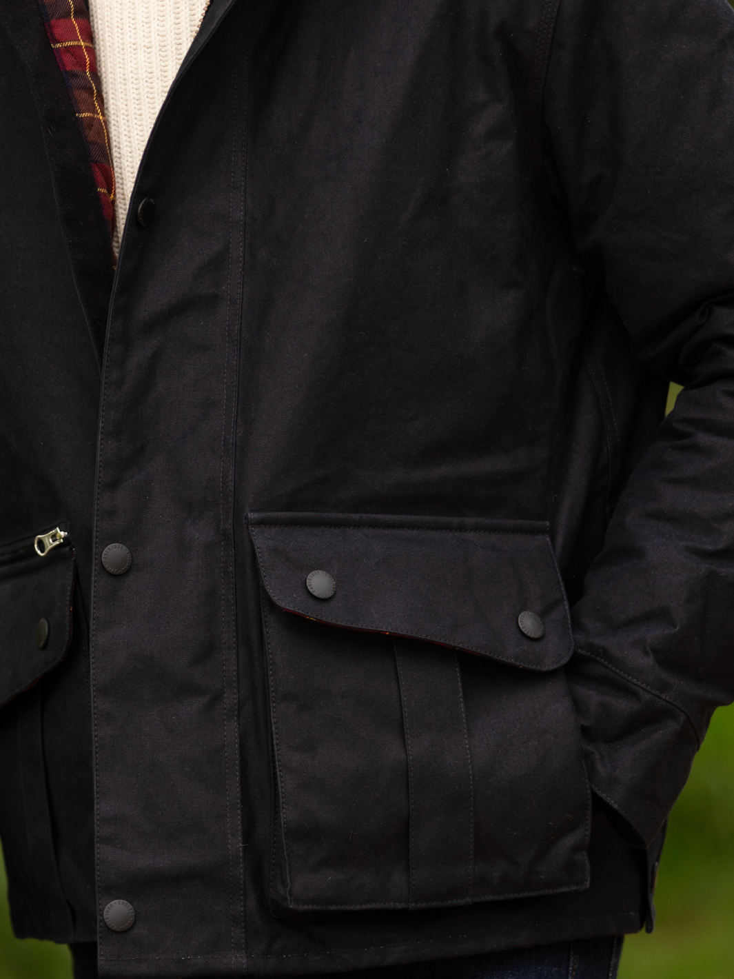 Hinterland Hybrid Jacket made of black dry waxed canvas with quilted shoulder and elbow patches, black moleskin trims, and red and burgundy check lining. Features include a hip-length design, oversized fit, action back, antique brass plate, and YKK zip with storm flap. Limited edition, part of the Partridge England range.