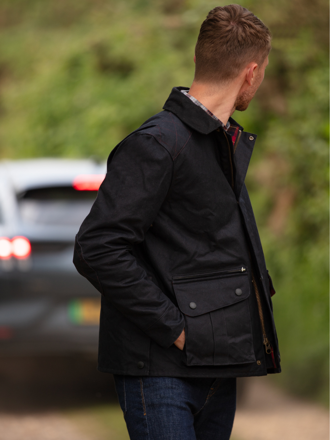 Hinterland Hybrid Jacket made of black dry waxed canvas with quilted shoulder and elbow patches, black moleskin trims, and red and burgundy check lining. Features include a hip-length design, oversized fit, action back, antique brass plate, and YKK zip with storm flap. Limited edition, part of the Partridge England range.