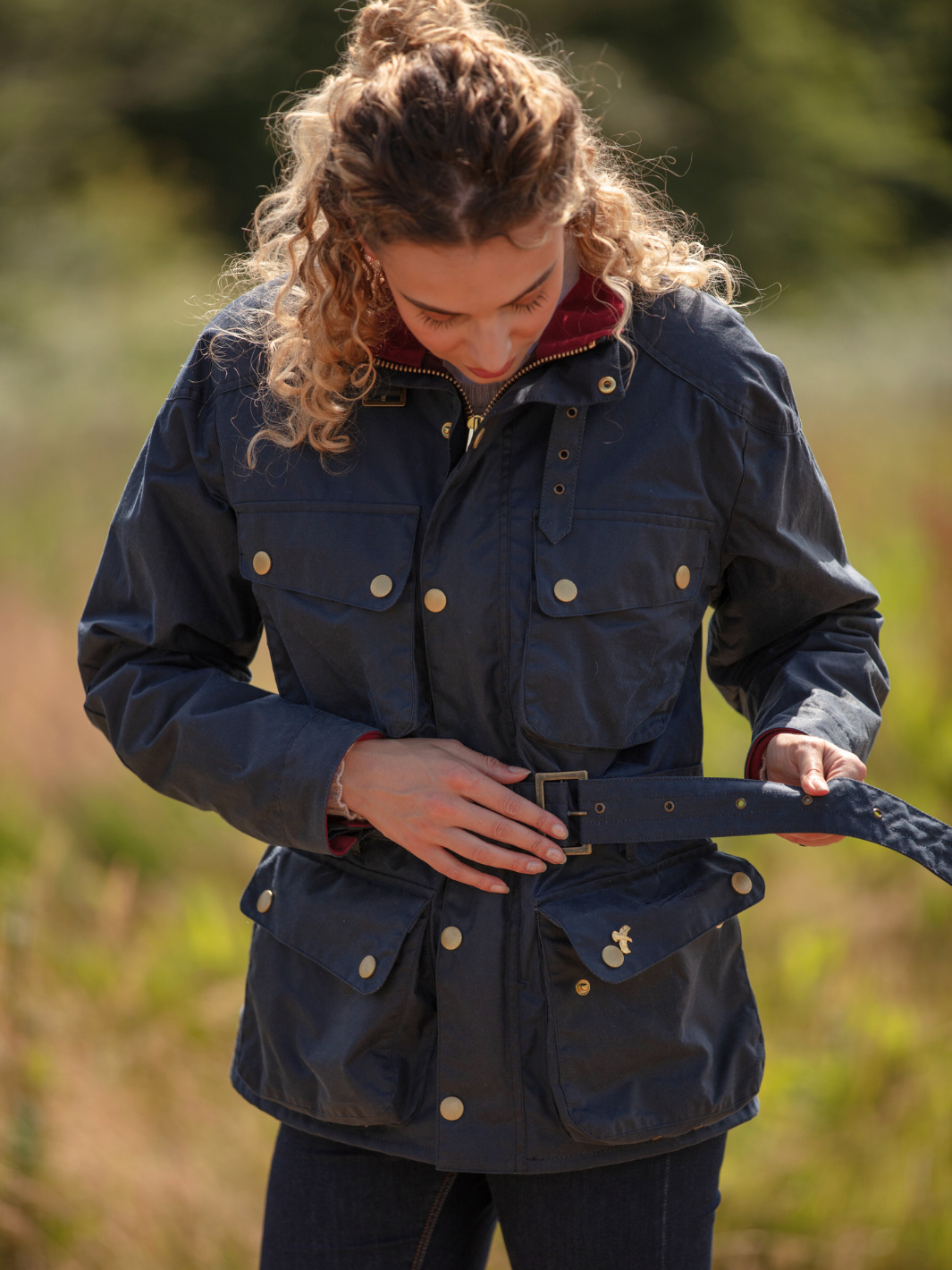 Wax Biker Jacket in Ocean Navy with cherry check lining, burgundy cotton moleskin collar, oversized pockets, and detachable belt. Crafted from British waxed material with tonal embroidery and gold-coloured studs for a blend of tradition, luxury, and modern function.