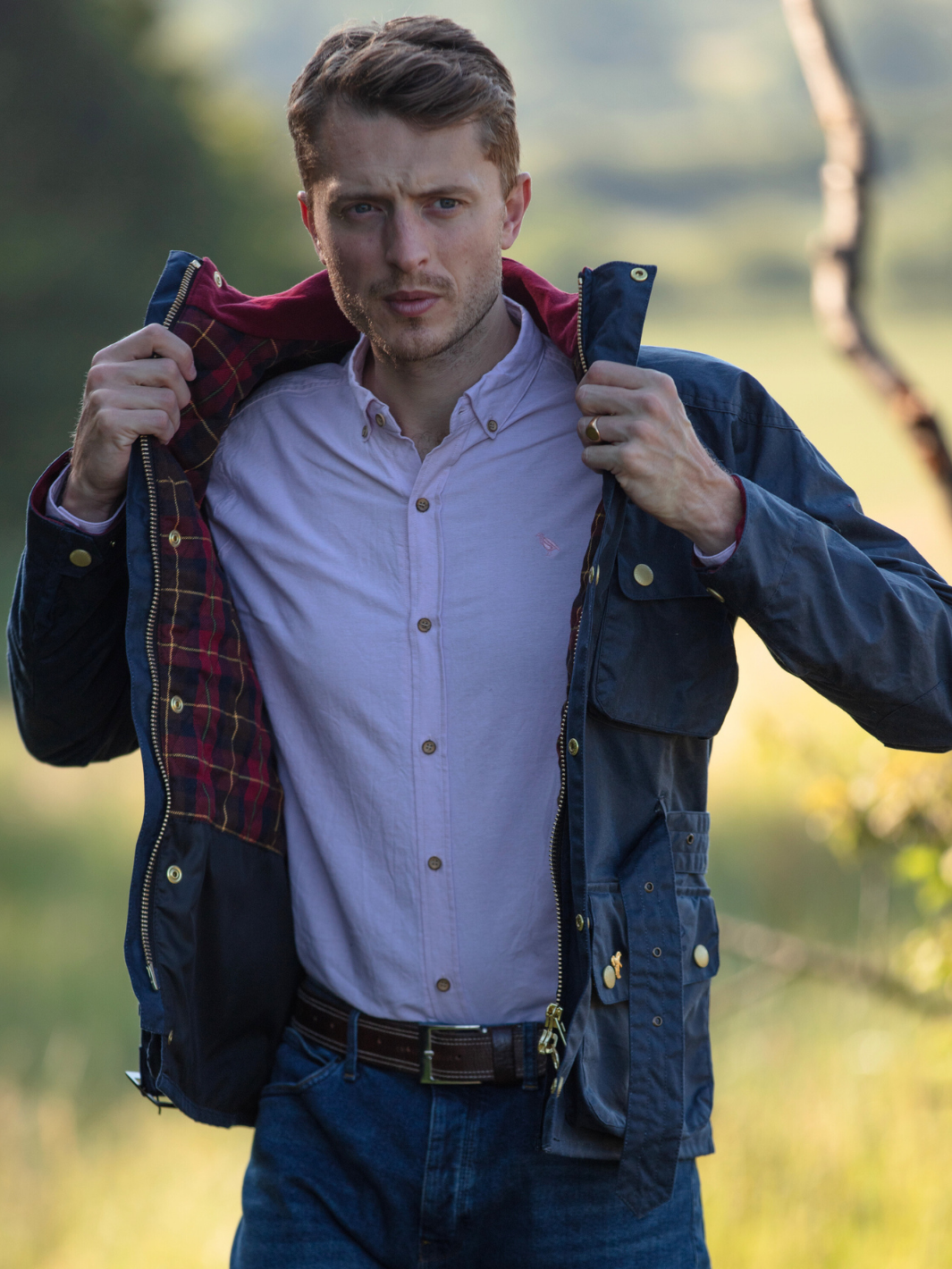 Men's Wax Biker Jacket in Ocean Navy with cherry check lining, burgundy cotton moleskin collar, oversized pockets, and detachable belt. Crafted from British waxed material with tonal embroidery and gold-coloured studs for a blend of tradition, luxury, and modern function.
